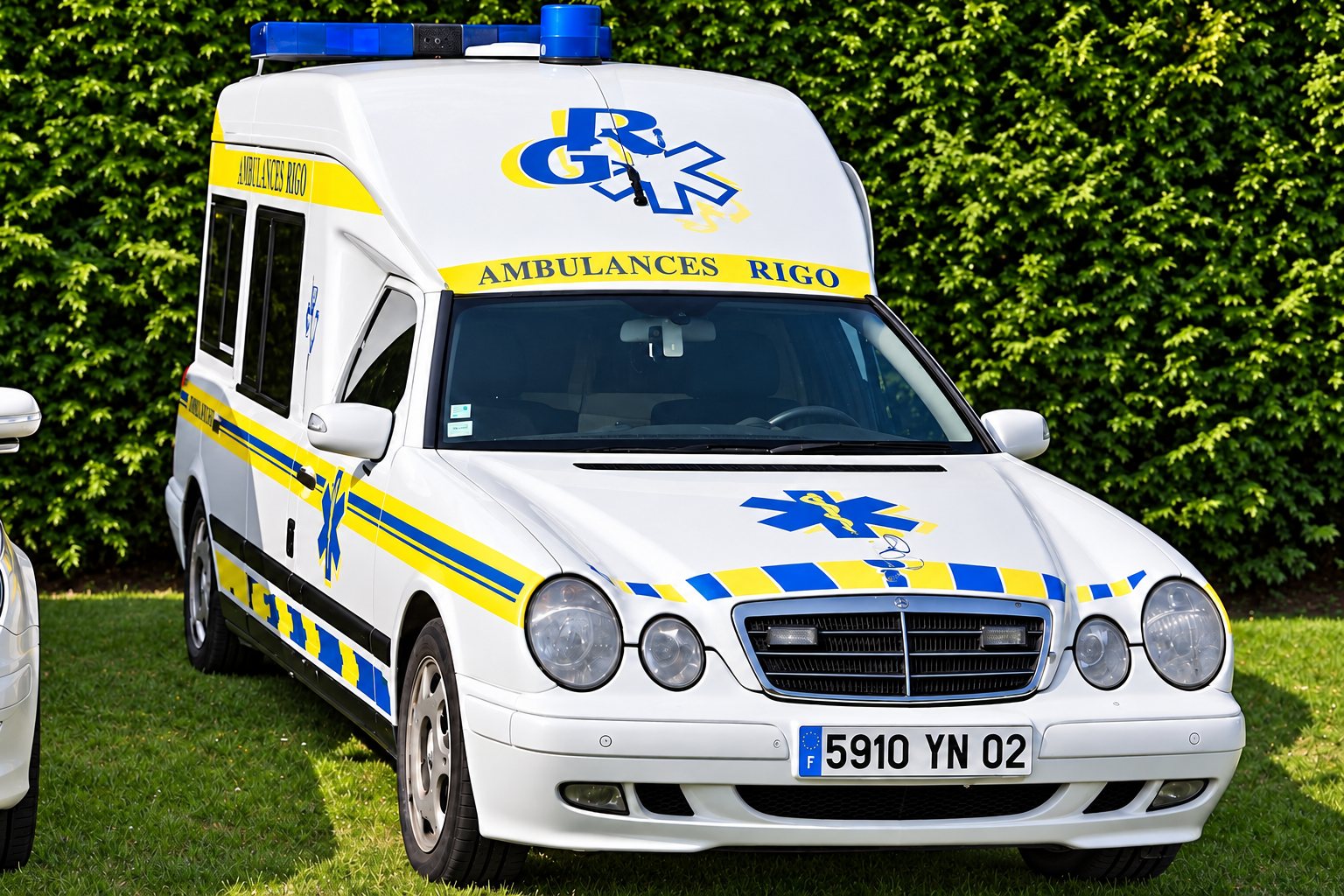 Ambulance blanche aux bandes jaunes et bleues, marquée Ambulances Rigo, vue de trois quarts avant sur une pelouse devant une haie, gyrophares bleus sur le toit.