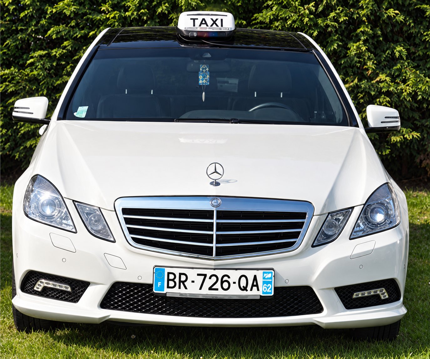 Berline blanche surmontée d’un lumineux Taxi, vue de face sur une pelouse devant une haie.